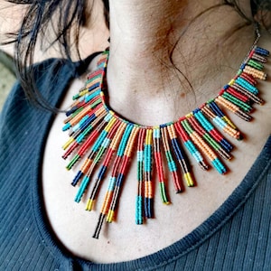 Può includere: Una collana di perline colorata con una vibrante gamma di colori tra cui rosso, blu, giallo e verde. La collana è composta da piccole perline cilindriche disposte in file verticali, creando un design accattivante. La collana è indossata al collo.