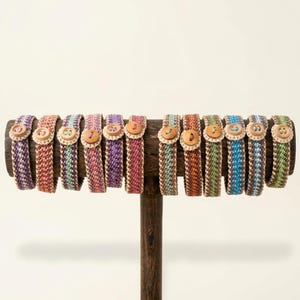 Puede incluir: Exhibición de pulseras tejidas en varios colores, incluyendo morado, rosa, verde y azul, cada una asegurada con un botón de madera. Las pulseras se muestran en un soporte de madera, mostrando sus intrincados patrones tejidos y tonos terrosos.