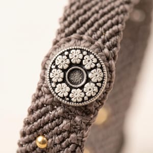 Könnte beinhalten: Ein taupefarbenes Makramee-Armband mit einem runden, silberfarbenen Knopfverschluss. Der Knopf ist mit einem Ring aus kleinen weißen Blumenmotiven verziert, die eine dunkle, kreisförmige Vertiefung in der Mitte umgeben. Kleine goldene Perlen sind sichtbar.