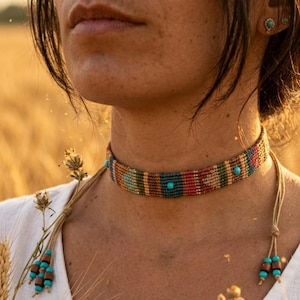 Puede incluir: Un collar tipo gargantilla tejido con un patrón geométrico multicolor en tonos azul, rojo, verde y beige. La banda está acentuada con pequeñas cuentas de color turquesa e incluye dos extremos de cordón colgantes, terminados con cuentas marrones y turquesas.