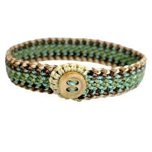 Peut inclure: Un bracelet fait à la main avec un motif tissé dans les tons de marron, vert et bleu. Il se ferme avec un bouton rond en bois clair. Le bracelet mesure environ 2,5 cm de large.