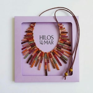 May include: A macrame necklace featuring a fringe of multicolored thread segments in shades of red, orange, brown, and green, displayed on a light purple card with the text HILOS DEL MAR. The necklace includes a brown cord with two wooden beads.