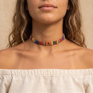 Puede incluir: Un collar tipo gargantilla tejido con un patrón geométrico repetitivo en colores vibrantes que incluyen rojo, naranja, amarillo, morado y azul. La banda está construida con múltiples hilos finos dispuestos horizontalmente para crear una superficie plana y texturizada.