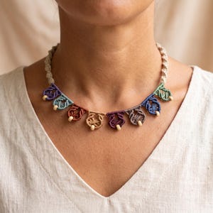 Puede incluir: Un collar tipo gargantilla de macramé con una base de cordón beige trenzado y una serie de pequeños colgantes anudados en varios colores, incluyendo morado, azul verdoso, óxido, beige, granate, marrón y azul, cada uno con una cuenta de madera.