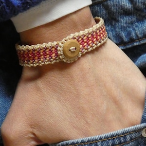 May include: A woven bracelet in beige, red, and orange hues, secured with a wooden button clasp. The bracelet is worn on a wrist, with a blue sweater and denim jeans in the background. The bracelet's design is intricate and detailed.