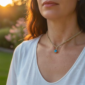Può includere: Una collana color oro con un pendente rotondo blu chiaro. La collana ha un design a catena intrecciata. La persona indossa un top bianco. Lo sfondo è sfocato con la luce del sole.
