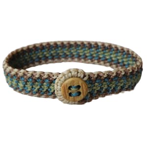 Può includere: Braccialetto fatto a mano con un motivo intrecciato nei toni del blu, verde e marrone. Il braccialetto è assicurato con un grande bottone rotondo in legno. Il design ricorda uno stile nautico o bohémien.