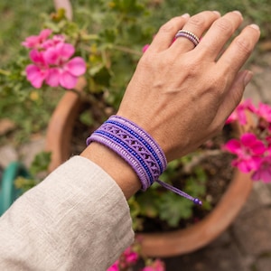 May include: A wide woven bracelet featuring alternating rows of light purple and dark blue beads with a central macrame pattern. The bracelet is secured with thin purple cords. A matching thin beaded ring is worn on the finger.