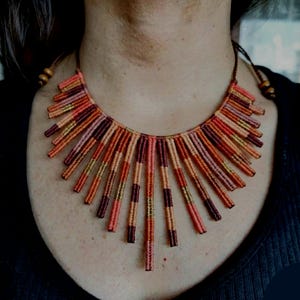 Puede incluir: Un collar hecho a mano con un diseño de sol. Cuenta con múltiples hebras verticales en tonos naranja, rojo y marrón, creando una apariencia vibrante y texturizada. El collar se lleva alrededor del cuello.