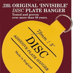 May include: A yellow round adhesive plate hanger with a 75mm diameter. The hanger has a loop at the top and the text "75mm / 3" DISC ADHESIVE PLATE HANGER Made in England" printed on it.
