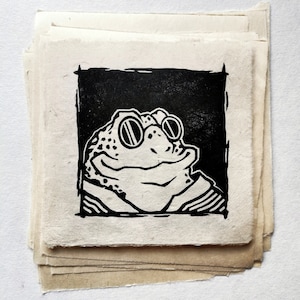 Peut inclure: Une pile d'estampes carrées en noir et blanc représentant une grenouille souriante portant des lunettes rondes. Les tirages sont réalisés sur du papier texturé.