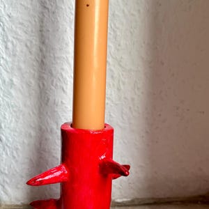 Puede incluir: Un portavelas rojo hecho a mano con un diseño único y texturizado. El soporte presenta varios elementos puntiagudos y salientes. Una vela beige se inserta en la parte superior. El portavelas es un artículo decorativo.