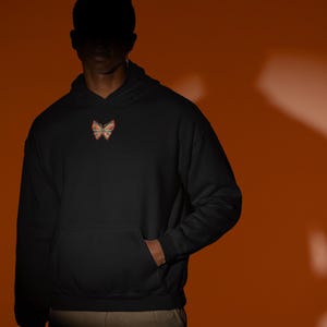 Puede incluir: Sudadera con capucha negra con un diseño de mariposa en el pecho. La mariposa es de color naranja, rosa y amarillo. La sudadera tiene un bolsillo delantero y una capucha con cordón.