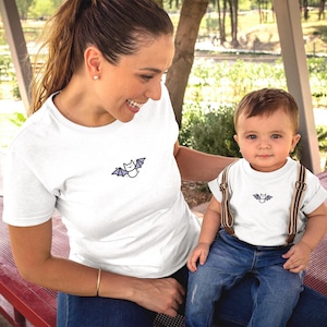 Puede incluir: Camisetas blancas con un diseño de murciélago. Una la lleva un adulto, la otra un niño. El niño lleva vaqueros azules y tirantes. El adulto lleva una pulsera dorada.