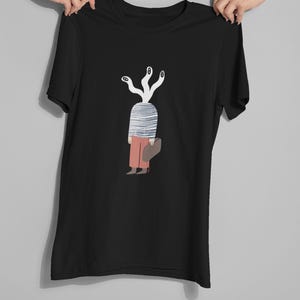 Puede incluir: Camiseta negra con una ilustración caprichosa. El diseño representa una figura con una cabeza a rayas y múltiples zarcillos blancos con forma de ojo. La figura lleva pantalones naranjas y lleva una bolsa marrón. La camiseta está sujeta por dos manos.