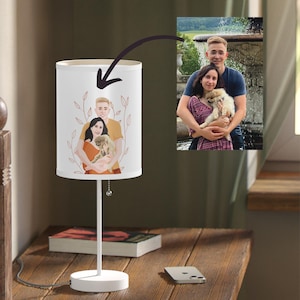 Puede incluir: Una lámpara de mesa blanca con una pantalla cilíndrica que presenta una ilustración personalizada de una pareja y un perro. La lámpara tiene una base de metal blanco y delgado y se encuentra sobre una superficie de madera. Un teléfono inteligente y libros también están sobre la mesa.