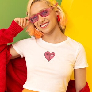 Puede incluir: Camiseta blanca de cuello redondo con un globo en forma de corazón rosa con una cara. La persona lleva gafas de sol rosas, pendientes rojos y una rebeca roja sobre una falda naranja. El fondo es verde y amarillo.