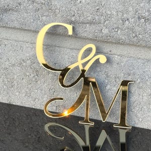 Może przedstawiać: Złoty monogram z literami "C" i "M" w eleganckiej, kursywnej czcionce. Litery są odblaskowe, z efektem lustrzanym widocznym poniżej. Tło to teksturowana, szara powierzchnia.