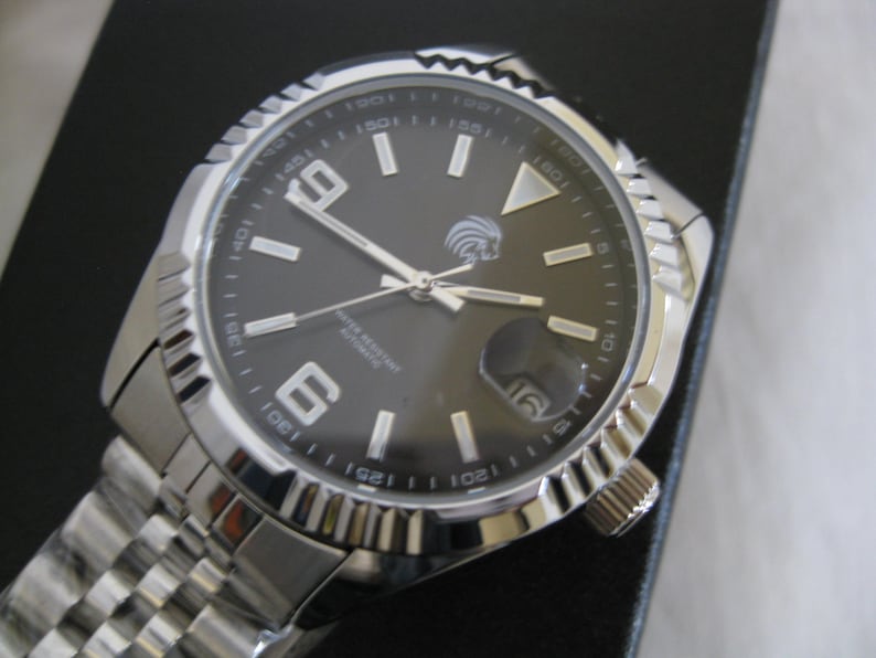 May include: A silver men's wristwatch with a black face and silver hands. The watch has a date window at the 3 o'clock position and a silver band.