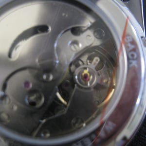 May include: Close-up view of the back of a silver watch with a clear case revealing the inner workings of the watch mechanism. The watch face has the word "BACK" printed on it.