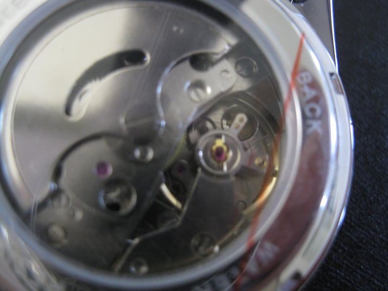 May include: Close-up view of the back of a silver watch with a clear case revealing the inner workings of the watch mechanism. The watch face has the word "BACK" printed on it.