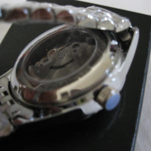 May include: A silver wristwatch with a stainless steel band and a silver face. The watch is on a black surface and the back of the watch is visible.
