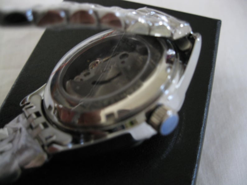 May include: A silver wristwatch with a stainless steel band and a silver face. The watch is on a black surface and the back of the watch is visible.