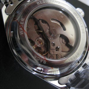 May include: Close-up of the back of a silver watch with a clear case showing the inner workings of the watch. The watch face says "WATER RESISTANT STAINLESS STEEL BACK".