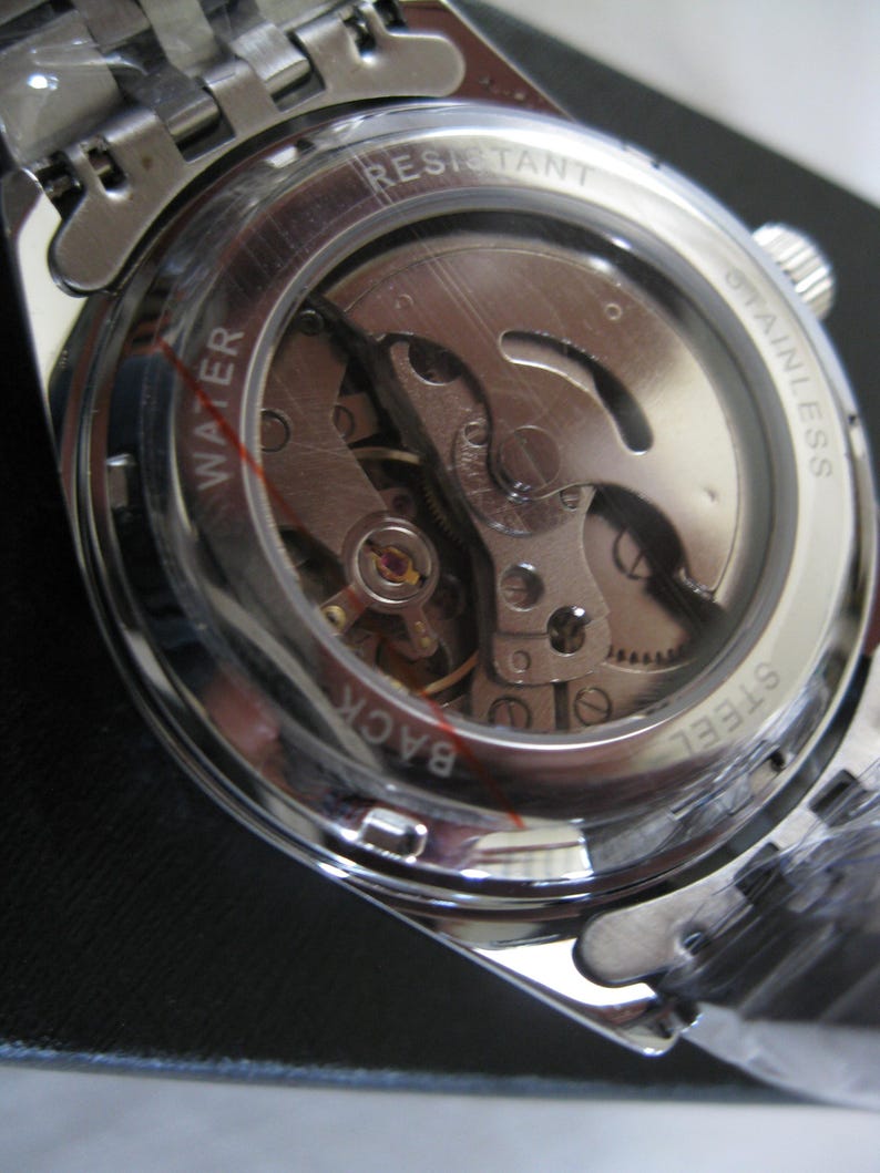 May include: Close-up of the back of a silver watch with a clear case showing the inner workings of the watch. The watch face says "WATER RESISTANT STAINLESS STEEL BACK".