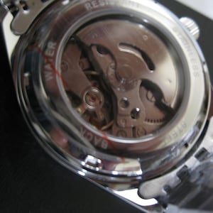 May include: A silver watch with a clear case back revealing the inner workings of the watch. The watch face has the words "WATER RESIST STAINLESS BACK" printed around the edge.