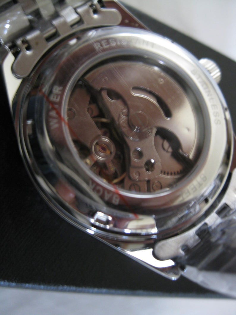 May include: A silver watch with a clear case back revealing the inner workings of the watch. The watch face has the words "WATER RESIST STAINLESS BACK" printed around the edge.