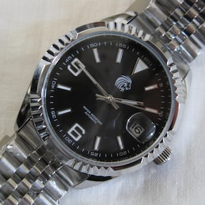 May include: A silver wristwatch with a black face and a silver metal band. The watch face has white markings and a date window at the 3 o'clock position. The watch face also has the text "WATER RESISTANT AUTOMATIC" printed on it.