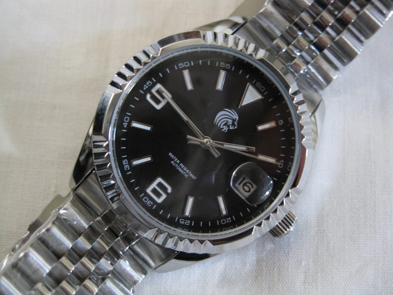 May include: A silver wristwatch with a black face and a silver metal band. The watch face has white markings and a date window at the 3 o'clock position. The watch face also has the text "WATER RESISTANT AUTOMATIC" printed on it.