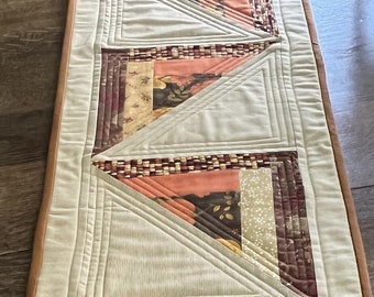 May include: A beige table runner with a brown border. The runner is made of fabric with a quilted design. The runner features a series of triangles made of different patterned fabrics in shades of brown, orange, and beige. A small wooden sign with the text "Hello Fall" is placed on the runner.