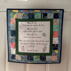 May include: A green and blue patchwork quilt with a white embroidered quote: "May the sun bring you new energy by day. May the moon softly restore you by night. May the rain wash away your worries. May you walk gently through the world and know it's beauty all the days of your life. apache blessing"