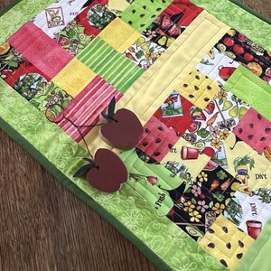 May include: A colorful quilted table runner with a patchwork design featuring red, green, and yellow squares. The runner is decorated with wooden apple accents. The border is a light green with a floral pattern.