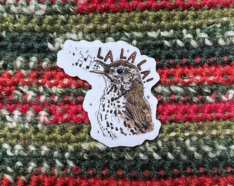 May include: A sticker depicting a brown and white bird singing "LA LA LA!" with musical notes. The sticker is set against a backdrop of red, green, and white knitted fabric. The bird is illustrated in a detailed style.