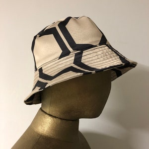 Puede incluir: Un sombrero de cubo beige con un patrón geométrico negro. El sombrero está hecho de una tela suave y sedosa.