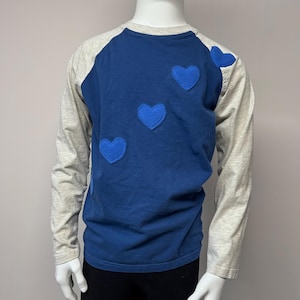 Puede incluir: Una camisa azul de manga larga con mangas grises y cuatro corazones de fieltro azul en la parte delantera.