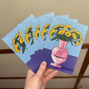 Puede incluir: Un conjunto de cinco tarjetas con una pintura de un ramo de girasoles amarillos en un jarrón rosa sobre un fondo azul. El jarrón está sobre una superficie morada.