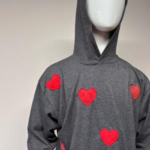 Puede incluir: Una sudadera con capucha gris con varios apliques de corazón rojo. La sudadera tiene una capucha y mangas largas. Los corazones están espaciados uniformemente en la parte delantera de la prenda, agregando un toque lúdico al estilo casual.