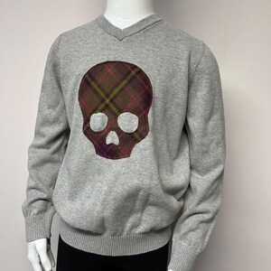 Puede incluir: Jersey gris de cuello en V con un gran diseño de calavera de cuadros en la parte delantera.