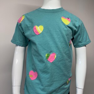 Puede incluir: Camiseta turquesa de manga corta con múltiples aplicaciones en forma de corazón. Los corazones presentan una combinación de colores rosa, amarillo y verde. La camiseta se muestra en un maniquí.