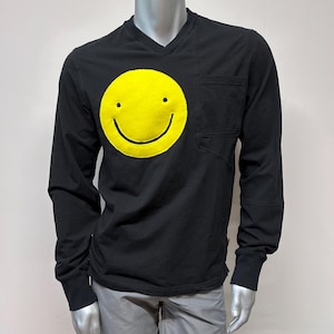 Puede incluir: Camiseta negra de manga larga con un gráfico de cara sonriente amarilla y un bolsillo en el lado izquierdo.