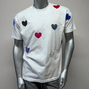 Puede incluir: Camiseta blanca con aplicaciones de corazones de colores. Los corazones son en tonos de rojo, azul, morado y gris.