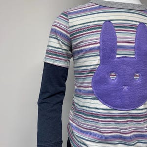 Puede incluir: Una camisa de rayas de manga larga con una aplicación de conejo de fieltro morado en la parte delantera. La camisa tiene una capa interior azul marino en las mangas.