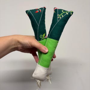 May include: Two stuffed fabric vegetable-shaped toys featuring a dark green top section with a red and white floral pattern, a bright lime green middle section, and a light beige bottom section with small white fringe threads at the base.
