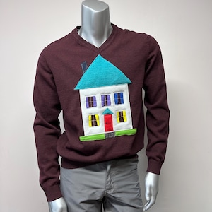 Puede incluir: Un suéter marrón con una aplicación de casa colorida. La casa tiene un techo turquesa, paredes blancas, ventanas amarillas y una puerta roja.