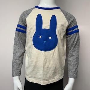 Puede incluir: Una camisa blanca de manga larga con mangas raglán azules y grises. La camisa tiene una aplicación de conejo de fieltro azul en la parte delantera.