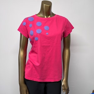 Puede incluir: Una camiseta rosa con lunares morados en el hombro izquierdo.